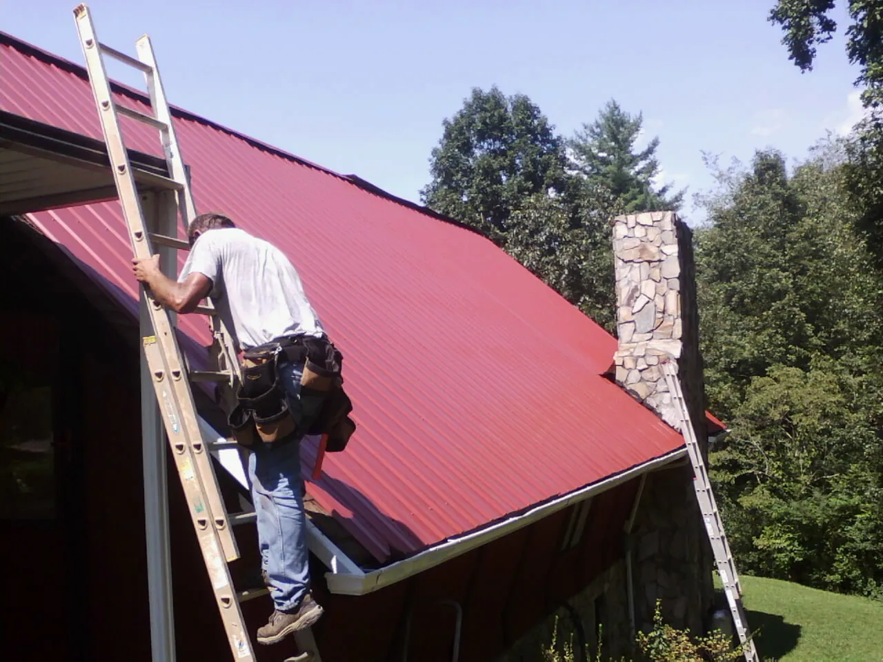 Roofer on a ladder beside a red metal roof and stone chimney — Soffit & Fascia Repair by Roofing Brothers in Winnebago